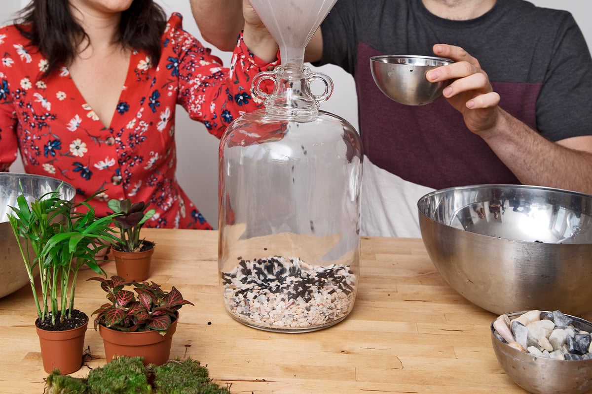 Twee personen vullen een fles met steentjes. Op de tafel staat verder een schaal met potgrond, plantjes en wat sierstenen.