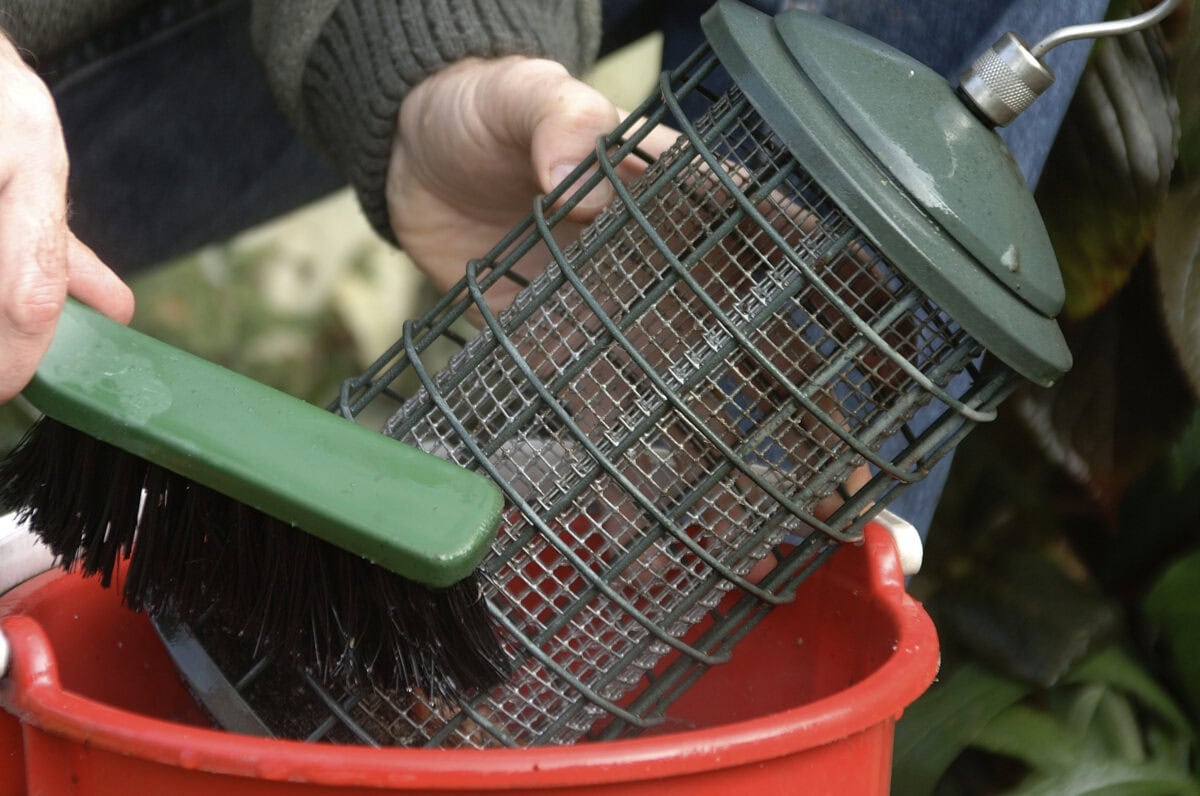 Iemand maakt met een borstel een vogelvoedersilo schoon in een sopje.
