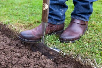 Twee laarzen staan achter een gereedschap waarmee graskanten worden afgestoken en er een nette rand ontstaat tussen kale grond en gazon.