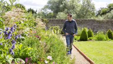 Adam Frost loopt over een recht tuinpad richting de kijker met aan de linkerkant een volle border die deels over het pad heen groeit.