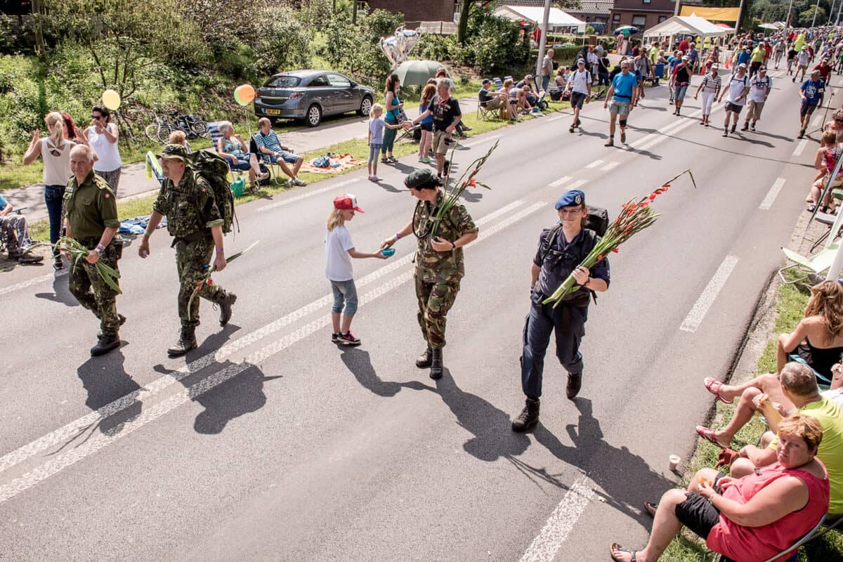 Vierdaagselopers op de weg, met toeschouwers aan de zijlijn. Militairen dragen gladiolen in hun armen en eentje krijgt er een.