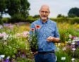 Marcel de Wagt met een plant in zijn hand, buiten op een veld met bloeiende planten.