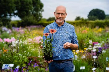 Marcel de Wagt met een plant in zijn hand, buiten op een veld met bloeiende planten.