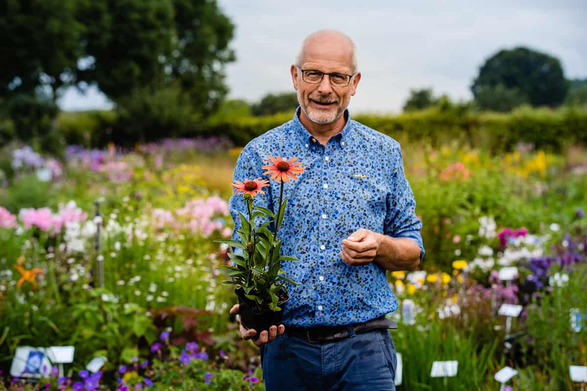 Marcel de Wagt met een plant in zijn hand, buiten op een veld met bloeiende planten.