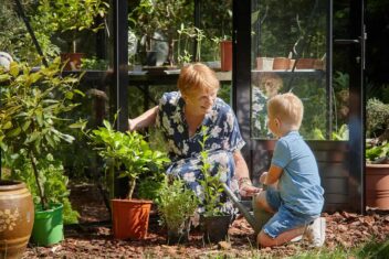 Een vrouw en een kind met potten vol planten met een serre vol potplanten op de achtergrond.