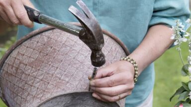 Twee handen houden een zinken teil vast en maken gaten in de onderkant met een hamer en spijker voor betere drainage.