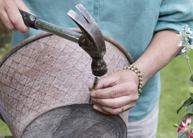 Twee handen houden een zinken teil vast en maken gaten in de onderkant met een hamer en spijker voor betere drainage.