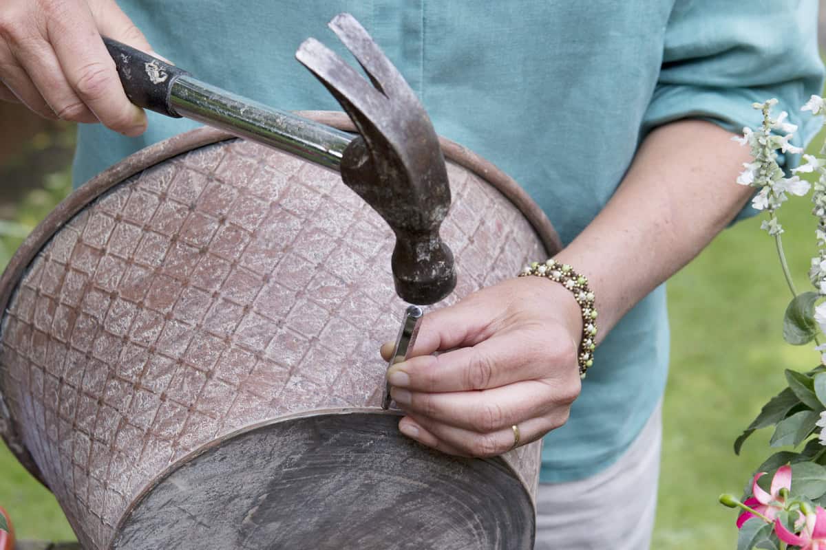 Twee handen houden een zinken teil vast en maken gaten in de onderkant met een hamer en spijker voor betere drainage.