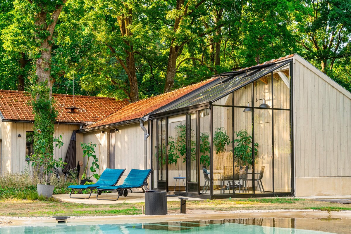Een serre, aangebouwd aan een verbouwde schuur, met twee ligstoelen en een zwembad ervoor.