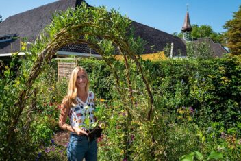 vrouw staat met bloemen in groene tuin voor gebouw.
