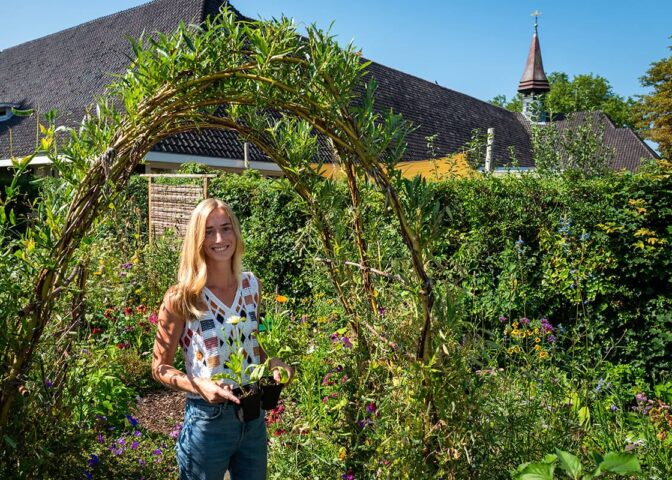 vrouw staat met bloemen in groene tuin voor gebouw.