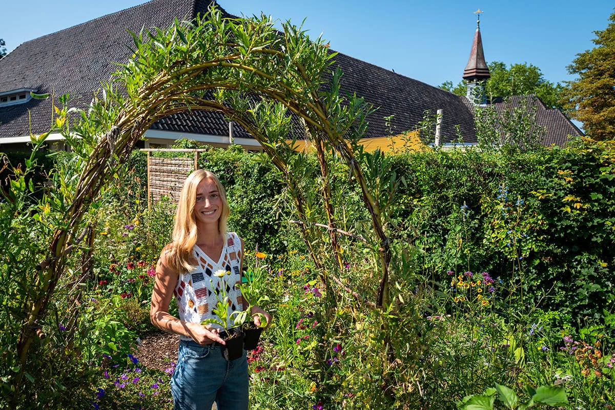 vrouw staat met bloemen in groene tuin voor gebouw.