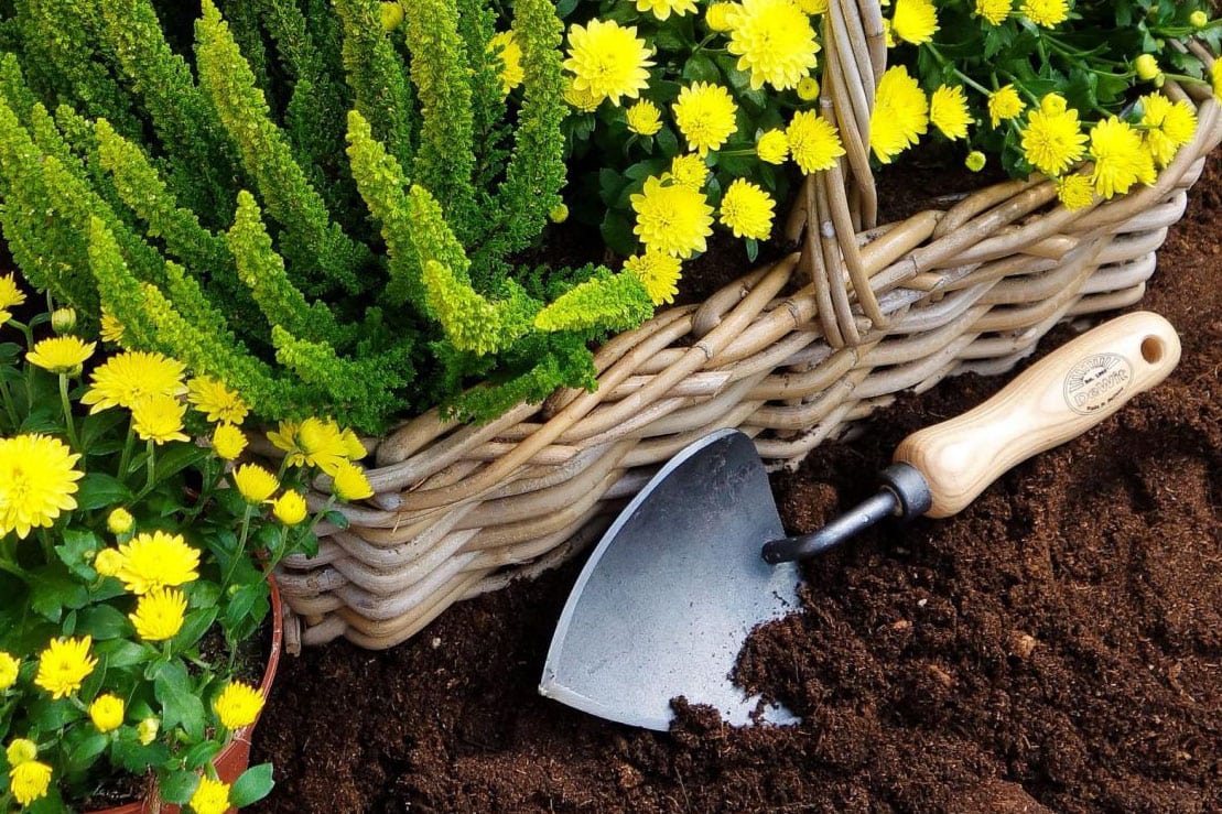 Een potschepje van De Wit ligt naast een mand met gele bloemen.