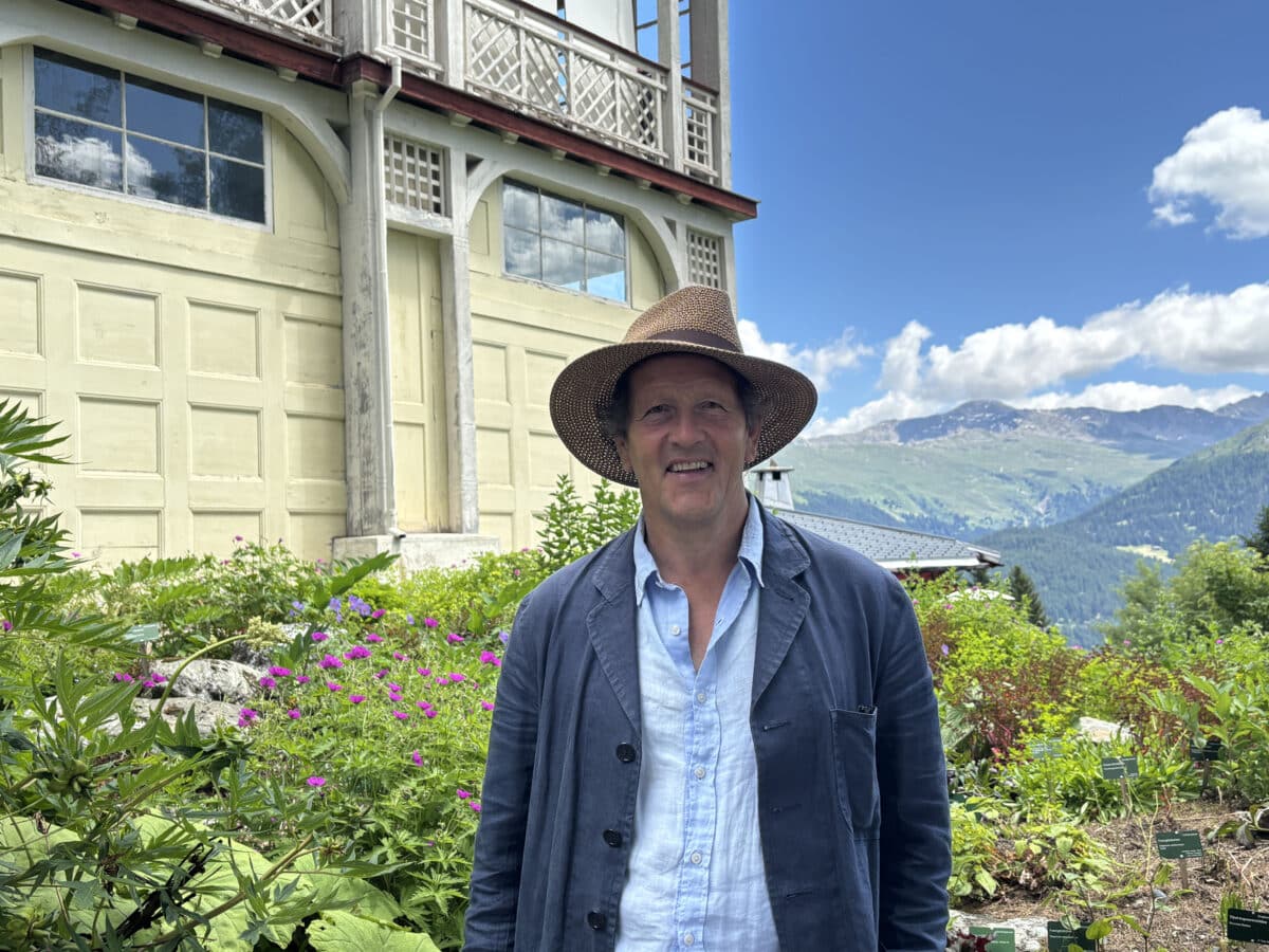 Monty Don in een tuin bij een groot landhuis en bergen op de achtergrond.