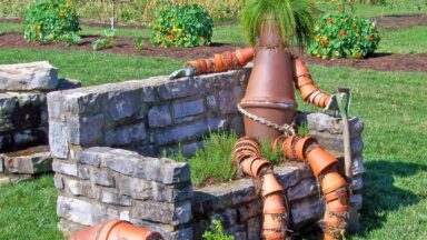Vogelverschrikker van potten en planten op een stenen bankje naast een hond van potten en planten.