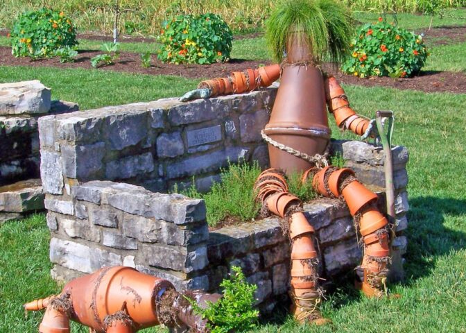 Vogelverschrikker van potten en planten op een stenen bankje naast een hond van potten en planten.