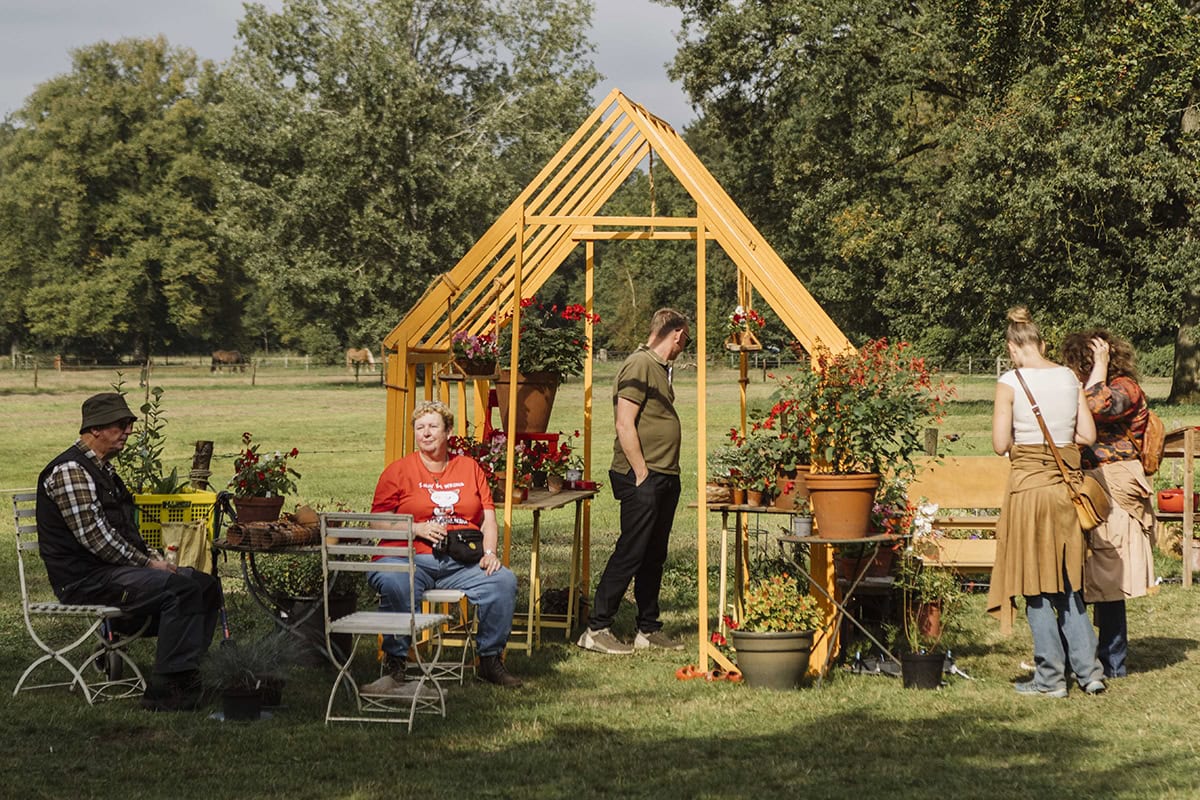 Gele kas met mensen eromheen die in het zonnetje zitten en lopen tussen de planten.