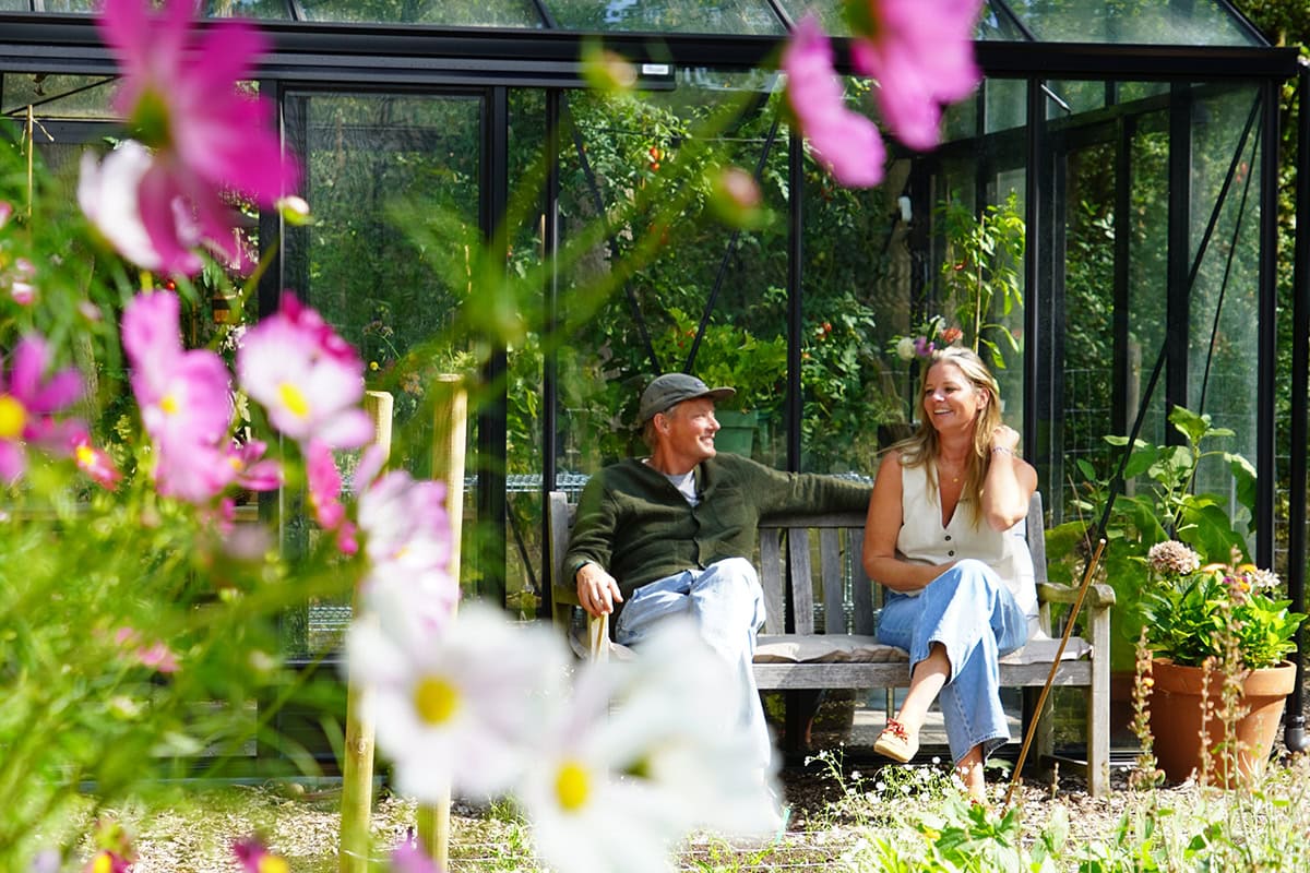 Twee mensen lachend op een bankje voor de kas met bloemen.