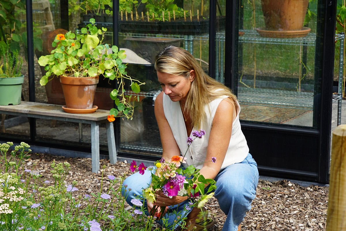 Vrouw zit gehurkt met bloemen in haar handen bij een kas.